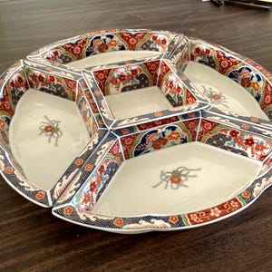 May include: A multi-compartment serving dish with a cream-colored interior and a decorative rim. The rim features a detailed pattern with red, blue, and green floral and geometric designs. The dish is likely made of ceramic.