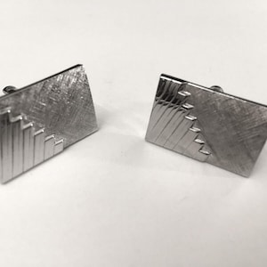 May include: A pair of silver-toned cufflinks. Each cufflink is rectangular with a textured surface and a stepped design on one corner. The cufflinks are set against a white background.
