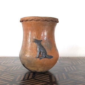 May include: A handcrafted, terracotta-colored vase with a textured surface and a dark gray dog illustration. The rim features a rope-like design. The vase is displayed on a dark wooden surface.