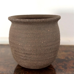 May include: A small, round, dark brown ceramic pot with a textured, matte surface. The vessel features a wide, slightly flared rim and subtle horizontal ridges along its body, showcasing a natural, earthy clay finish with visible speckles.