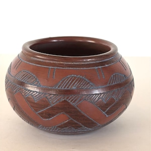 May include: A round, terracotta-colored ceramic pot with a wide opening. The pot features a carved geometric design in a darker brown and blue. The design includes horizontal lines and angular shapes, creating a textured appearance. The pot is likely handmade.