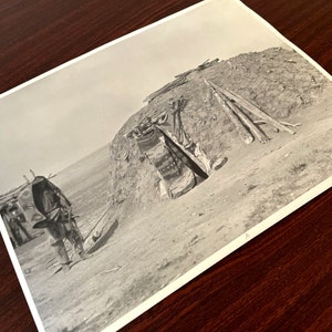 May include: A black and white photograph of a person standing in front of a dirt structure. The person is wearing a long coat and a hat. The structure is a dome-shaped building with a small opening at the top.