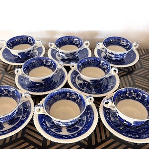 May include: A set of eight blue and white porcelain teacups and saucers. The cups have two handles and are decorated with a floral and landscape design. The saucers have a scalloped edge. The set is arranged on a dark wooden surface.