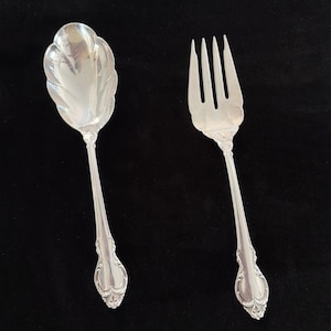May include: A silver spoon and fork set against a black background. The spoon has a scalloped bowl and a decorative handle. The fork has four tines and a matching handle. Both pieces are ornate and likely part of a formal dining set.