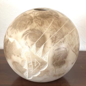 May include: A spherical vase with a smooth, matte finish in shades of cream and light brown. The surface features a marbled, cloud-like pattern and a single, deeply carved geometric design consisting of sharp angles and lines on the front.