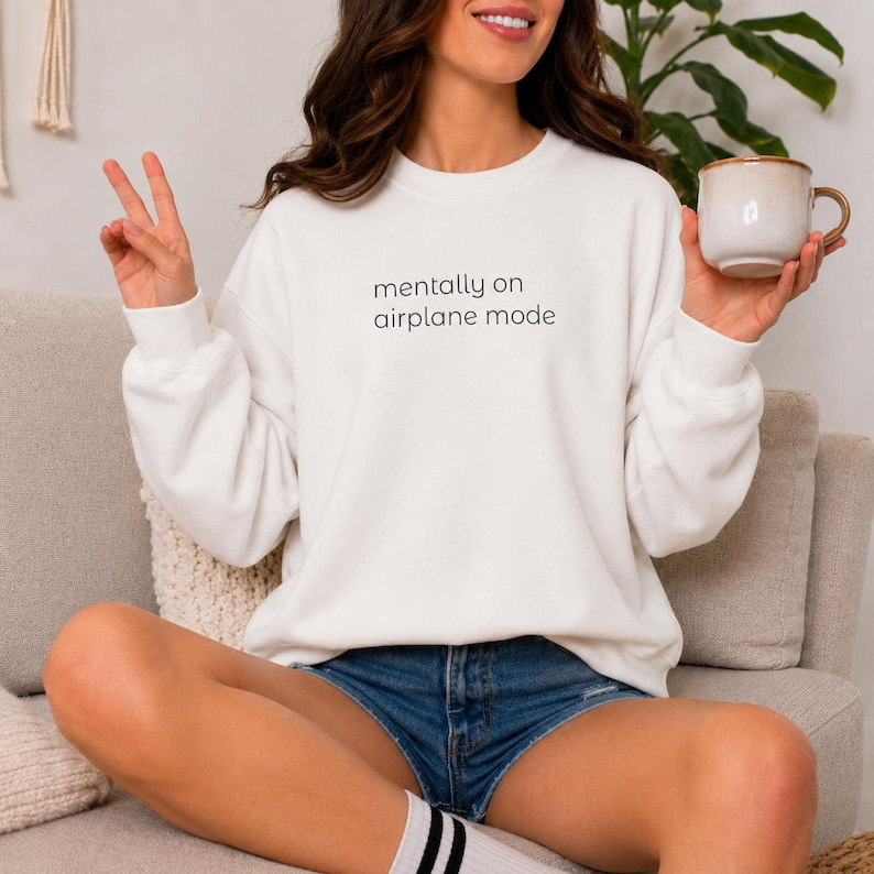 Puede incluir: Una sudadera blanca de cuello redondo con el texto "mentally on airplane mode" en negro. La sudadera se combina con pantalones cortos de mezclilla. Una persona sostiene una taza beige con un asa marr&oacute;n.