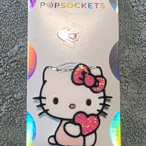 May include: A white Hello Kitty PopSockets phone grip featuring a glittery pink bow and a glittery pink heart. The character has black whiskers, black eyes, and a small pink nose. The product is mounted on a silver-toned backing card with the text POPSOCKETS.