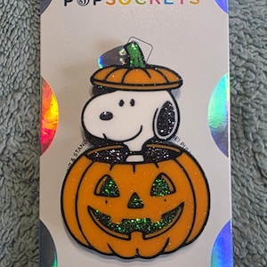 May include: A PopSockets PopGrip featuring a white Snoopy character with black ears sitting inside an orange glittery jack-o'-lantern pumpkin with a matching orange pumpkin lid on top. The product is mounted on a white backing card with holographic circles.