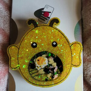 May include: A yellow glitter bee-shaped phone grip with a clear, round, glittery center filled with colorful confetti and small bee charms. The grip is labeled "POPGRIP" and "POPSOCKETS" on the packaging.