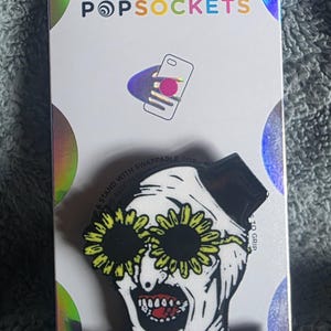 May include: A PopSockets phone grip featuring a graphic of a clown with white face paint, a black collar, and yellow sunflower-shaped sunglasses. The clown has a wide, open-mouthed expression with visible teeth. The item is attached to its original packaging.