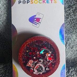 May include: A circular phone grip on a white backing card labeled POPSOCKETS and POPGRIP. The grip features a red glitter border and a clear center filled with black glitter and small horror-themed charms, including a masked figure and a hand.