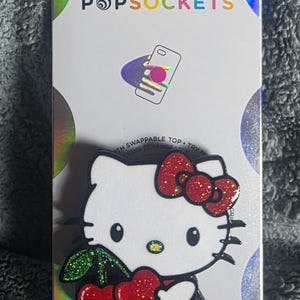 May include: A white PopSockets phone grip featuring a Hello Kitty character design with a red glittery bow, a red polka dot dress, and holding two red glittery cherries with green leaves, mounted on a white backing card with holographic accents.