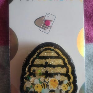 May include: A black and gold PopSocket PopGrip with a beehive design. The beehive is filled with glitter and small bee and flower charms. The text "PopGrip" is printed in pink, blue, and black letters.