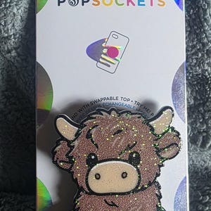 May include: A PopSockets phone grip featuring a glittery, cartoon-style brown Highland cow with small horns, black hooves, and a light tan muzzle. The product is mounted on a white retail display card with the text POPSOCKETS and a graphic illustration.