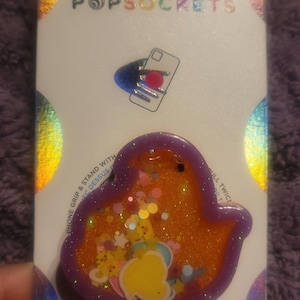 May include: A purple and orange PopSocket phone grip with a glittery, translucent design. The grip features a yellow duck with a pink heart and stars. The packaging says "PopGrip" and "Be Rooted".
