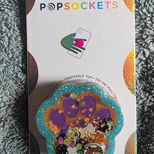 May include: A paw-shaped phone grip on a white backing card with the text POPSOCKETS. The grip features a teal glitter border, a purple paw pad, and a clear resin center filled with colorful confetti, including small bone shapes and cartoon character images.