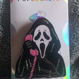 May include: A black and white glittery PopGrip phone accessory featuring a character in a hooded robe holding a pink telephone receiver. The product is mounted on a white backing card with the text POPSOCKETS and POPGRIP printed in colorful letters.