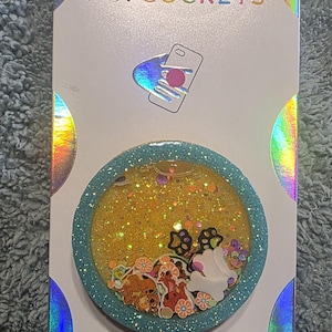 May include: A circular phone grip on a white backing card with holographic accents. The grip features a teal glitter border and a yellow liquid-filled center containing small decorative shapes, including paw prints, bones, and cartoon characters.