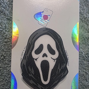 May include: A black and white Ghostface mask phone grip on a holographic backing card. The card features the text POPSOCKETS at the top and POPGRIP at the bottom. The mask design is centered on the card, showcasing a white face with black eyes and a wide open mouth, surrounded by a black hooded cloak.