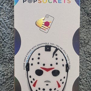 May include: A white PopSockets phone grip and stand featuring a hockey mask design with black dots, red accents, and a black outline. The product is mounted on a white cardboard backing with the text POPSOCKETS and instructions to pull twice to grip.