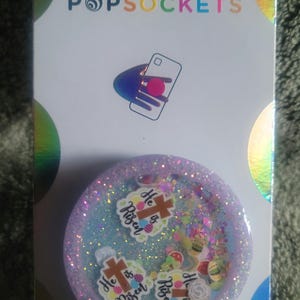May include: A light blue and white glitter phone grip with a cross and the words "He is Risen" in pink and white lettering. The grip is on a white card with the words "PopSockets" and "PopGrip" printed on it.