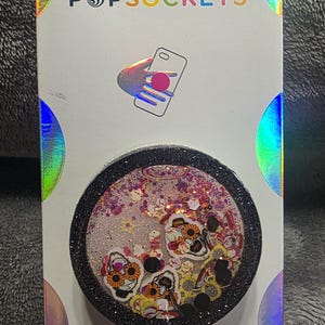 May include: A circular phone grip with a black glittery rim and a clear liquid-filled center containing floating skull figures with sunflower accents, pink glitter, and small stars. The product is mounted on a white card labeled POPSOCKETS and POPGRIP.