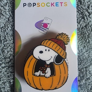 May include: A PopSockets PopGrip phone accessory featuring a graphic of Snoopy wearing a red and yellow knit hat and scarf while sitting inside a large orange pumpkin. The product is mounted on a white backing card with holographic accents.