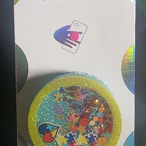 May include: A circular PopSockets phone grip with a bright yellow glitter border and a clear center filled with colorful floating confetti, including puzzle piece shapes and a heart, mounted on a white retail display card with branding.