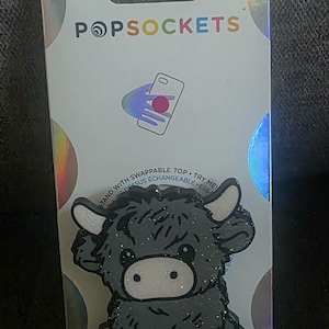 May include: A PopSockets phone grip featuring a dark gray, glittery Highland cow design with white horns and a white muzzle. The product is attached to its original white retail packaging, which displays the brand name PopSockets and a graphic illustration of the grip in use.