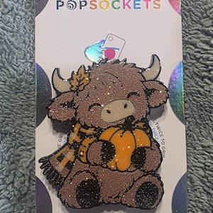 May include: A PopSockets PopGrip featuring a glittery brown Highland cow illustration wearing a yellow and black plaid scarf and holding an orange pumpkin. The cow has a small leaf on its head. The packaging displays the text PopSockets and PopGrip.
