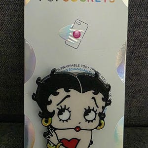 May include: A PopSockets phone grip featuring a Betty Boop character design with black hair, gold hoop earrings, and a red heart graphic on the clothing. The item is mounted on a white retail display card with the text POPSOCKETS at the top.