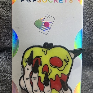 May include: A PopSockets phone grip featuring a graphic of a red apple covered in dripping neon yellow slime, held by a pale skeletal hand with long black fingernails. The item is mounted on a white branded display card with holographic accents.