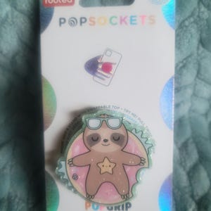 May include: A green and pink PopSocket phone grip with a sloth wearing sunglasses and a pink donut. The text "GRIP & STAND WITH SWAPPABLE TOP - TRY ME! PULL TWICE" is printed on the grip. The text "POPSOCKETS" is printed on the packaging.