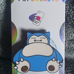 May include: A Snorlax character PopGrip phone accessory mounted on a white backing card with holographic accents. The PopGrip features a blue and cream-colored Snorlax graphic. The packaging displays the text POPSOCKETS and POPGRIP.
