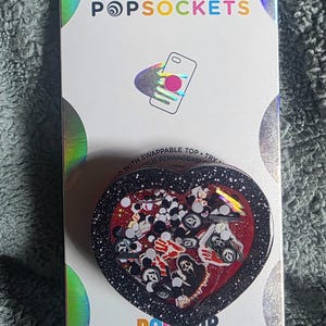 May include: A heart-shaped PopSockets PopGrip on a white backing card with holographic accents. The grip features a black glitter border and a red interior filled with small, loose horror-themed icons, including ghost-like masks and red handprints.