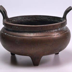 May include: A bronze-colored, three-legged incense burner with two arched handles. The vessel has a rounded body and a wide, open top. The metal shows signs of age and wear, with a slightly mottled appearance.