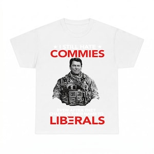Może przedstawiać: Biały t-shirt z krótkim rękawem z czarno-białą grafiką przedstawiającą Ronalda Reagana w sprzęcie taktycznym. Tekst brzmi I STILL HATE COMMIES na górze oraz EVEN AFTER THEY CHANGED THEIR NAME TO LIBERALS na dole w kolorze czerwonym i białym.