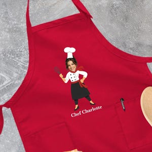 May include: A bright red apron featuring a graphic illustration of a chef wearing a white hat and uniform, holding a whisk. The text "Chef Charlotte" is printed in white below the figure. The apron includes a front pocket and adjustable neck strap.