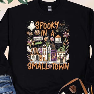 May include: A black long-sleeve sweatshirt featuring a colorful graphic print of a small town with ghosts, a witch, and flowers. The text reads "SPOOKY IN A SMALL TOWN" with the number "689883" displayed on a sign above the illustrated buildings.