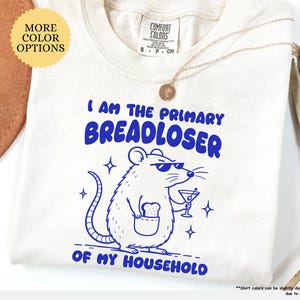 May include: An ivory short-sleeved t-shirt featuring a blue graphic of a rat wearing sunglasses, holding a martini glass, and carrying a slice of bread in its pocket. The text reads I AM THE PRIMARY BREADLOSER OF MY HOUSEHOLD in blue letters.