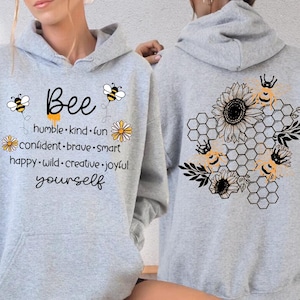 May include: A sport grey hooded sweatshirt featuring a front graphic with the text Bee humble, kind, fun, confident, brave, smart, happy, wild, creative, joyful, yourself and two bees. The back displays a black honeycomb pattern with sunflowers and bees.