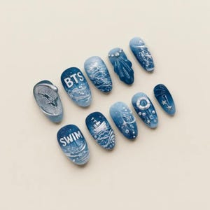 May include: A set of ten press-on nails featuring an ocean theme with blue gradient colors. Designs include the text BTS and SWIM, a shark, a ship, a lifebuoy, stars, and bubbles. The nails have a glossy finish with raised 3D details and accents.