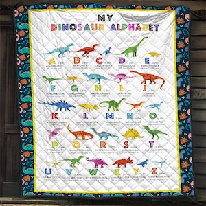 May include: A white quilted blanket featuring the text MY DINOSAUR ALPHABET with colorful illustrations of dinosaurs corresponding to each letter from A to Z. The border is dark blue with a repeating pattern of small dinosaurs and leaves.