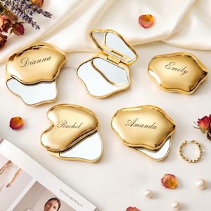 May include: Four gold-colored, irregular-shaped compact mirrors with smooth, reflective surfaces. Each mirror is engraved with a name: Dianna, Rachel, Amanda, and Emily. One mirror is shown open, revealing the interior glass, while the others are closed.