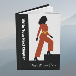 May include: A hardcover book with a black spine and white cover featuring a silhouette of a person in an orange jumpsuit with a yellow floral belt, stepping up a black block. The text reads Write Your Next Chapter and Your Name Here.