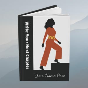 May include: A hardcover book with a black spine and white cover. The spine reads "Write Your Next Chapter" in white text. The cover features a silhouette of a person in an orange outfit with a yellow floral belt, stepping onto a black block.