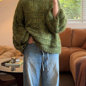 Puede incluir: Un jersey de punto verde oliva de gran tamaño con cuello redondo y mangas anchas. El jersey se combina con vaqueros holgados de color azul claro. El conjunto es informal y cómodo.