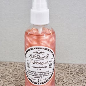 May include: A clear plastic spray bottle containing a shimmering peach-coloured shower and body oil. The label reads 2Lip BeautEh, Bubblegum, 1 oz, and lists ingredients including grapeseed, almond, coconut, and jojoba oils with vitamin E.