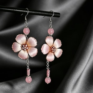 Può includere: Orecchini pendenti con grandi pendenti a forma di fiore rosa chiaro. Ogni orecchino ha una catena d'argento con una perla rosa e un accento a righe rosa e bianco. Gli orecchini sono sospesi a una barra nera su uno sfondo scuro.