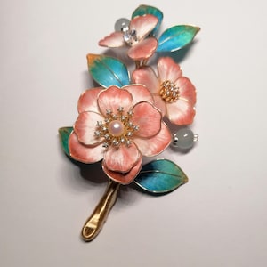 May include: A floral brooch with pink and white flowers, gold detailing, and teal and green leaves. The brooch has a pearl centre and is embellished with crystals. The stem is gold. A decorative piece of jewellery.